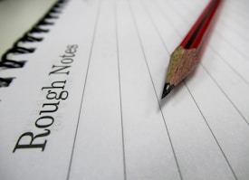 A wooden pencil with a red and black design lies on a lined notebook page. The text 'Rough Notes' is printed on the left side of the notebook. The image conveys a sense of simplicity and focus on writing or note-taking.