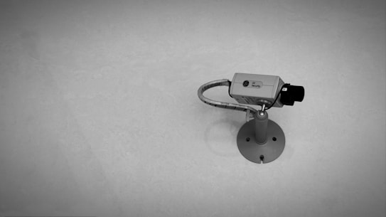 A wall-mounted security camera is positioned on a sturdy base with a protective casing and a flexible conduit for the wiring. The background is a smooth, plain surface.