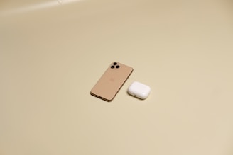 A gold smartphone and a white wireless earbud case sit on a plain beige surface. The devices are positioned with a sense of balance, the smartphone features a triple-camera setup at the back.