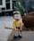 A cartoon figurine with large, round glasses and a big smile is holding a baseball bat. The figurine is dressed in a yellow shirt and blue shorts with white socks and blue shoes. The background is a blurry outdoor setting with some greenery and a stone.