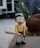A cartoon figurine with large, round glasses and a big smile is holding a baseball bat. The figurine is dressed in a yellow shirt and blue shorts with white socks and blue shoes. The background is a blurry outdoor setting with some greenery and a stone.