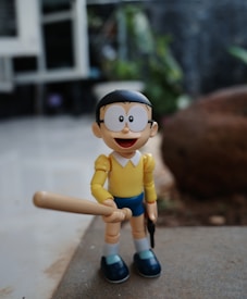 A cartoon figurine with large, round glasses and a big smile is holding a baseball bat. The figurine is dressed in a yellow shirt and blue shorts with white socks and blue shoes. The background is a blurry outdoor setting with some greenery and a stone.