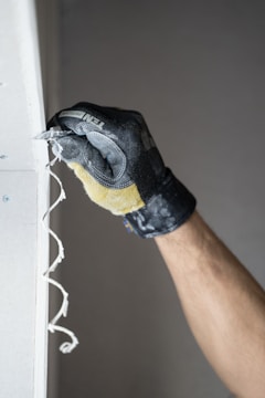 A gloved hand is scraping along the edge of a wall, creating a spiraled shaving of material. The glove is black with a textured surface, and the wall is light in color with visible screws.