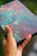 A hand holding a colorful abstract painting with vibrant swirls of red, green, blue, and yellow on a rectangular canvas.