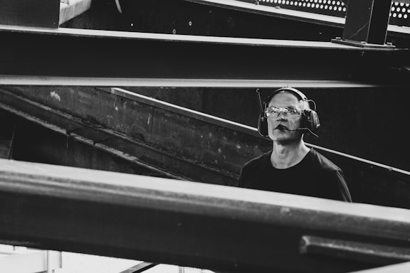 A man wearing protective eyewear and earmuffs appears to be working or observing within an industrial or construction site. Steel beams and metal structures dominate the scene, creating an atmosphere of heavy machinery and engineering.