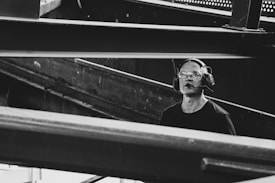 A man wearing protective eyewear and earmuffs appears to be working or observing within an industrial or construction site. Steel beams and metal structures dominate the scene, creating an atmosphere of heavy machinery and engineering.