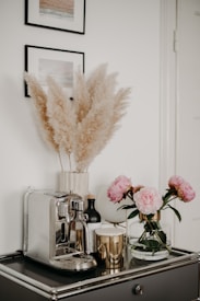 A stylish kitchen or living space corner features a sleek silver espresso machine on a dark, glossy countertop. Accompanying the machine are several decorative items, including a gold metallic candle, a black bottle, and a glass vase holding pink peonies. Above the counter on the wall are two framed pictures, and a vase of pampas grass adds a touch of softness to the arrangement.