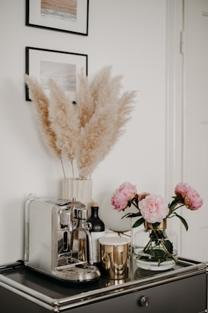 A stylish kitchen or living space corner features a sleek silver espresso machine on a dark, glossy countertop. Accompanying the machine are several decorative items, including a gold metallic candle, a black bottle, and a glass vase holding pink peonies. Above the counter on the wall are two framed pictures, and a vase of pampas grass adds a touch of softness to the arrangement.