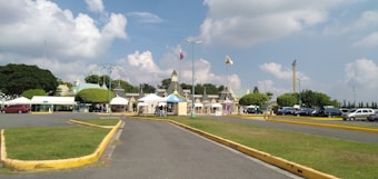 A paved road leads to a grand entrance surrounded by lush green grass and neatly trimmed trees. Several colorful structures with pointed roofs and flags create a festive atmosphere. In the background, a Ferris wheel can be seen against a backdrop of fluffy white clouds and a clear blue sky. Cars are parked along the road, adding to the busy and vibrant scene.