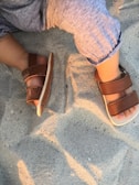 A child's feet wearing brown sandals are seen resting on soft sand, with the child dressed in light blue pants.