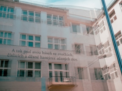 The image features the reflection of a building with multiple windows and a modern architectural design. Overlaid on the reflection is a Czech text, creating a layered visual effect.
