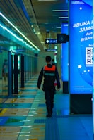 A security officer is walking down a modern, brightly lit hallway with shiny tiles and digital displays. The person is wearing a dark uniform with a reflective vest. The corridor seems to be part of a transportation hub or metro station with directional signs overhead.