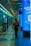 A security officer is walking down a modern, brightly lit hallway with shiny tiles and digital displays. The person is wearing a dark uniform with a reflective vest. The corridor seems to be part of a transportation hub or metro station with directional signs overhead.