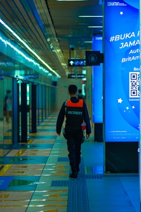 A security officer is walking down a modern, brightly lit hallway with shiny tiles and digital displays. The person is wearing a dark uniform with a reflective vest. The corridor seems to be part of a transportation hub or metro station with directional signs overhead.