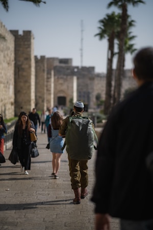 A group of people walking along a stone-paved path near an ancient stone wall, with a person in military attire and a large backpack in the foreground. Palm trees are visible along the path, and distant buildings are in the background.