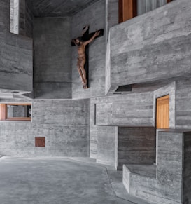 A modern, minimalist interior made of exposed concrete surfaces with geometric architectural features. A wooden crucifix is mounted on one of the walls, adding a spiritual focal point to the otherwise stark environment. There are a few small wooden and glass windows recessed into the concrete walls, offering a contrast in texture and color.