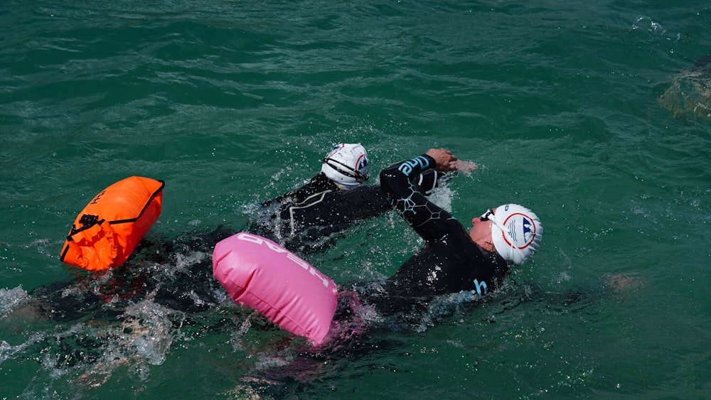 2 person in black and white wet suit on pink inflatable float