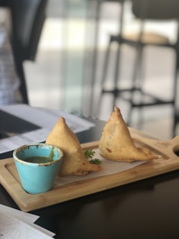 Two triangular fried pastries are presented on a wooden serving board beside a small, blue bowl filled with a green dipping sauce. The pastries are likely samosas, and a sprig of fresh coriander is placed next to them.