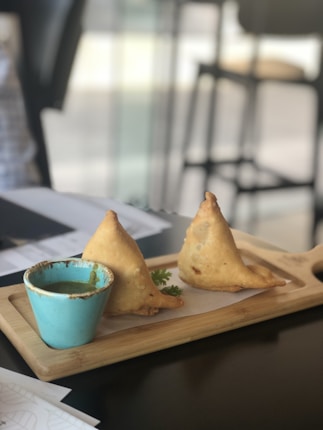 Two triangular fried pastries are presented on a wooden serving board beside a small, blue bowl filled with a green dipping sauce. The pastries are likely samosas, and a sprig of fresh coriander is placed next to them.