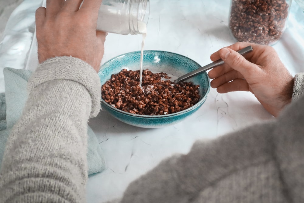 person holding stainless steel spoon and white ceramic bowl with brown food