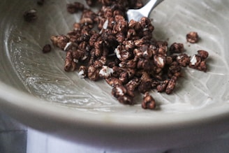 A ceramic bowl contains a small portion of chocolate-covered puffed cereal. The cereal pieces are irregularly shaped, with a glossy coating and white flecks. A metal spoon is partially visible, resting in the bowl, suggesting readiness for eating.