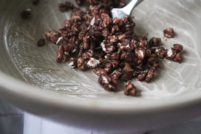 A ceramic bowl contains a small portion of chocolate-covered puffed cereal. The cereal pieces are irregularly shaped, with a glossy coating and white flecks. A metal spoon is partially visible, resting in the bowl, suggesting readiness for eating.