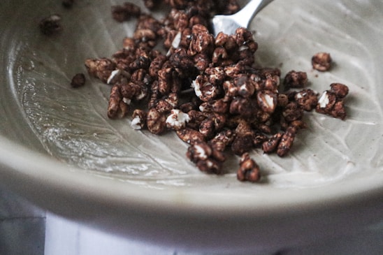 A ceramic bowl contains a small portion of chocolate-covered puffed cereal. The cereal pieces are irregularly shaped, with a glossy coating and white flecks. A metal spoon is partially visible, resting in the bowl, suggesting readiness for eating.