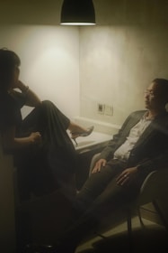 Two people seated in a dimly lit room, with a single overhead light casting a warm glow. One person, dressed in a dark outfit, has their legs up on a seat and appears relaxed. The other person, wearing a suit, sits across from them with an attentive demeanor. The setting suggests a casual or intimate conversation in an otherwise plain environment.