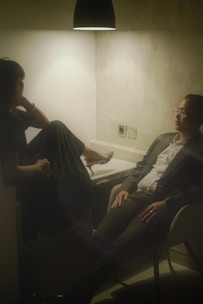 Two people seated in a dimly lit room, with a single overhead light casting a warm glow. One person, dressed in a dark outfit, has their legs up on a seat and appears relaxed. The other person, wearing a suit, sits across from them with an attentive demeanor. The setting suggests a casual or intimate conversation in an otherwise plain environment.