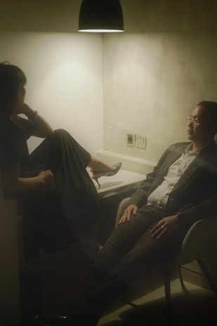 Two people seated in a dimly lit room, with a single overhead light casting a warm glow. One person, dressed in a dark outfit, has their legs up on a seat and appears relaxed. The other person, wearing a suit, sits across from them with an attentive demeanor. The setting suggests a casual or intimate conversation in an otherwise plain environment.