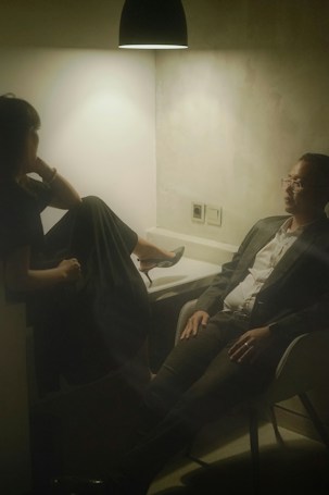 Two people seated in a dimly lit room, with a single overhead light casting a warm glow. One person, dressed in a dark outfit, has their legs up on a seat and appears relaxed. The other person, wearing a suit, sits across from them with an attentive demeanor. The setting suggests a casual or intimate conversation in an otherwise plain environment.