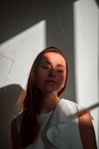 A person with closed eyes stands in soft light, casting artistic shadows on a plain background. Sunlight creates geometric patterns around the face and shoulders, enhancing the serene expression. The person is wearing a white garment with textured details.
