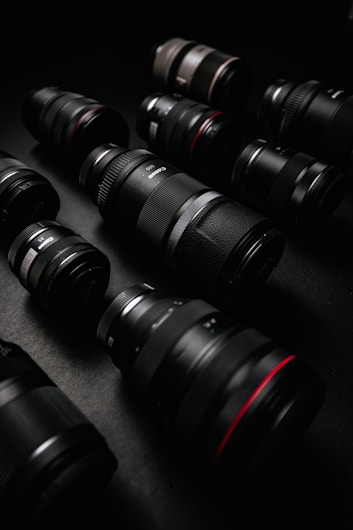 A collection of camera lenses is arranged on a dark surface. The lenses vary in size and appear to be made of black material with some red accents. The lighting highlights the texture and details of the lenses.