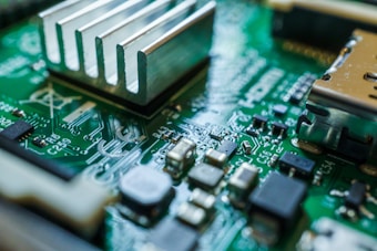 A close-up view of a green circuit board featuring various electronic components such as capacitors, resistors, and a heat sink. The intricate design of the circuitry can be seen with silver solder traces and black components set against the green base material.