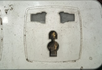 A close-up view of a dirty electrical wall socket with visible grime and stains. The socket design is for a plug with two flat and one round pin. The surrounding area shows signs of wear and lack of maintenance.