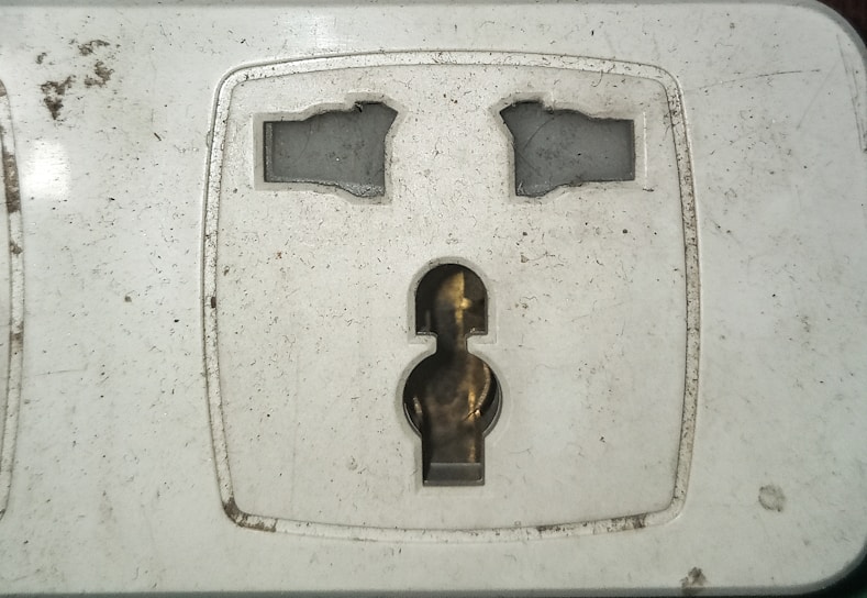 A close-up view of a dirty electrical wall socket with visible grime and stains. The socket design is for a plug with two flat and one round pin. The surrounding area shows signs of wear and lack of maintenance.