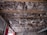 The ceiling of a room under construction or renovation, with visible wooden beams and exposed fiberglass insulation. Loose and fluffy pink insulation material is tucked between the beams, with some electrical wires hanging down. The walls are partially uncovered, showing exposed bricks and some unfinished drywall.