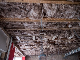The ceiling of a room under construction or renovation, with visible wooden beams and exposed fiberglass insulation. Loose and fluffy pink insulation material is tucked between the beams, with some electrical wires hanging down. The walls are partially uncovered, showing exposed bricks and some unfinished drywall.