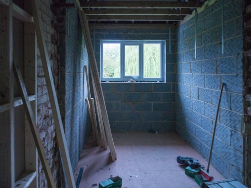 A partially constructed room with exposed brick walls and concrete blocks. Several wooden planks are leaning against the left wall. The window in the far wall allows natural light to illuminate the space. Tools and construction materials are scattered on the plywood floor.