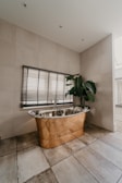 A luxurious bathroom features a copper freestanding bathtub positioned against a tiled wall, accompanied by Venetian blinds on the window above. A large green potted plant adds a touch of nature, while the earthy tones of the floor tiles complement the metallic sheen of the bathtub.