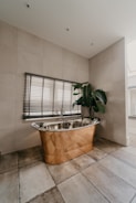 A luxurious bathroom features a copper freestanding bathtub positioned against a tiled wall, accompanied by Venetian blinds on the window above. A large green potted plant adds a touch of nature, while the earthy tones of the floor tiles complement the metallic sheen of the bathtub.