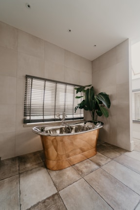 A luxurious bathroom features a copper freestanding bathtub positioned against a tiled wall, accompanied by Venetian blinds on the window above. A large green potted plant adds a touch of nature, while the earthy tones of the floor tiles complement the metallic sheen of the bathtub.