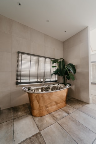 A luxurious bathroom features a copper freestanding bathtub positioned against a tiled wall, accompanied by Venetian blinds on the window above. A large green potted plant adds a touch of nature, while the earthy tones of the floor tiles complement the metallic sheen of the bathtub.