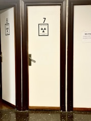 A row of closed doors numbered 7 and 8 with radiation warning signs attached. The signs indicate a monitored area and external irradiation risk. The doors are framed in dark wood, creating a contrast with the light-colored walls. The floor is made of dark, reflective tiles.