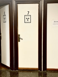 A row of closed doors numbered 7 and 8 with radiation warning signs attached. The signs indicate a monitored area and external irradiation risk. The doors are framed in dark wood, creating a contrast with the light-colored walls. The floor is made of dark, reflective tiles.