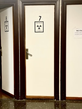 A row of closed doors numbered 7 and 8 with radiation warning signs attached. The signs indicate a monitored area and external irradiation risk. The doors are framed in dark wood, creating a contrast with the light-colored walls. The floor is made of dark, reflective tiles.