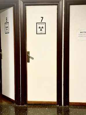 A row of closed doors numbered 7 and 8 with radiation warning signs attached. The signs indicate a monitored area and external irradiation risk. The doors are framed in dark wood, creating a contrast with the light-colored walls. The floor is made of dark, reflective tiles.