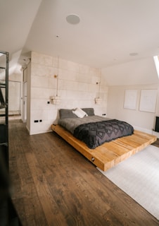 A modern bedroom with a minimalist design featuring a low wooden platform bed. The bedding consists of dark gray and white pillows, set against a textured white stone wall. Two pendant lights hang from the ceiling, adding a touch of elegance. The room includes light wooden flooring and simple line art on the walls. Natural light enters from a window, creating a warm, serene atmosphere.