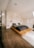 A modern bedroom with a minimalist design featuring a low wooden platform bed. The bedding consists of dark gray and white pillows, set against a textured white stone wall. Two pendant lights hang from the ceiling, adding a touch of elegance. The room includes light wooden flooring and simple line art on the walls. Natural light enters from a window, creating a warm, serene atmosphere.