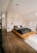 A modern bedroom with a minimalist design featuring a low wooden platform bed. The bedding consists of dark gray and white pillows, set against a textured white stone wall. Two pendant lights hang from the ceiling, adding a touch of elegance. The room includes light wooden flooring and simple line art on the walls. Natural light enters from a window, creating a warm, serene atmosphere.